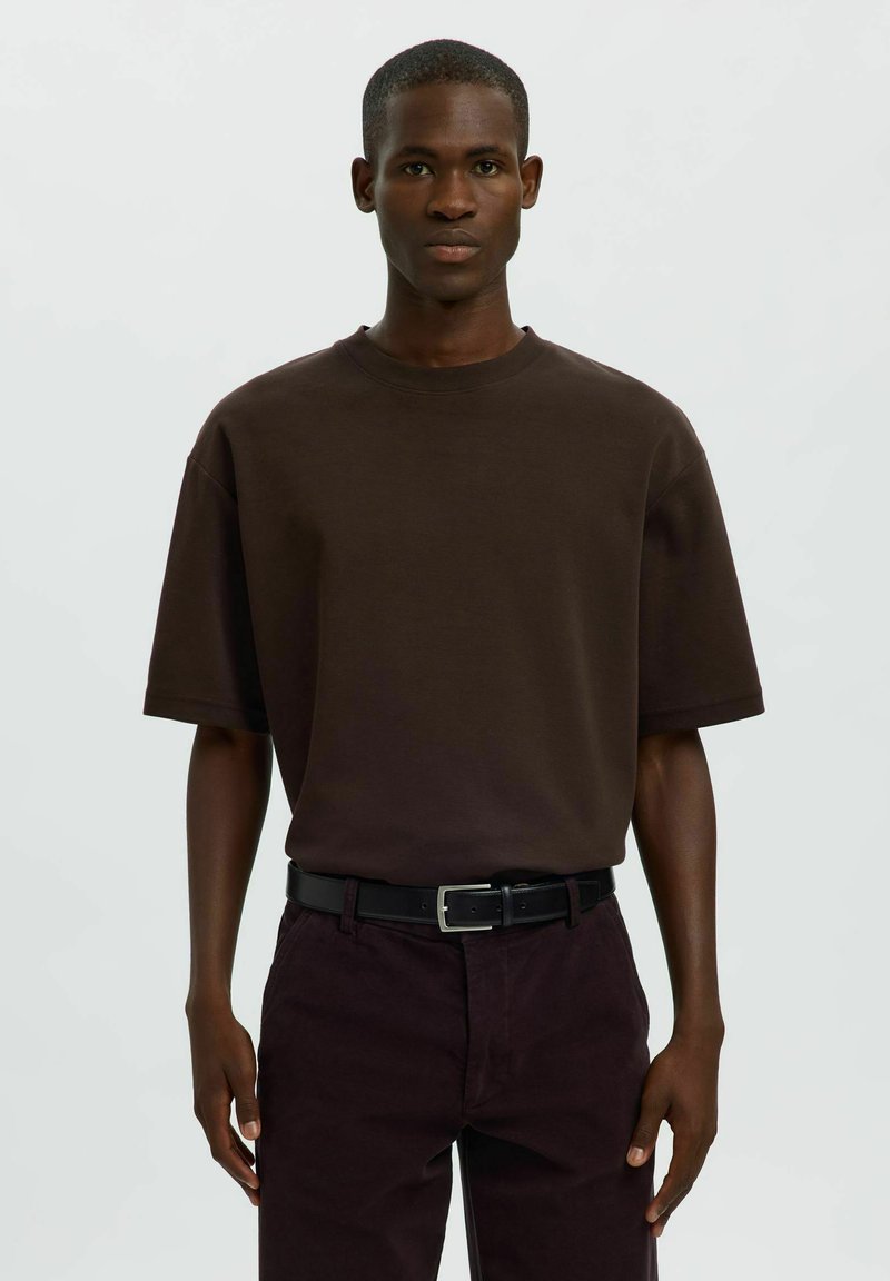 Jeune homme noir portant une chemise marron foncé oversize à manches courtes, une ceinture noire et un pantalon foncé, debout devant un fond clair uni.
