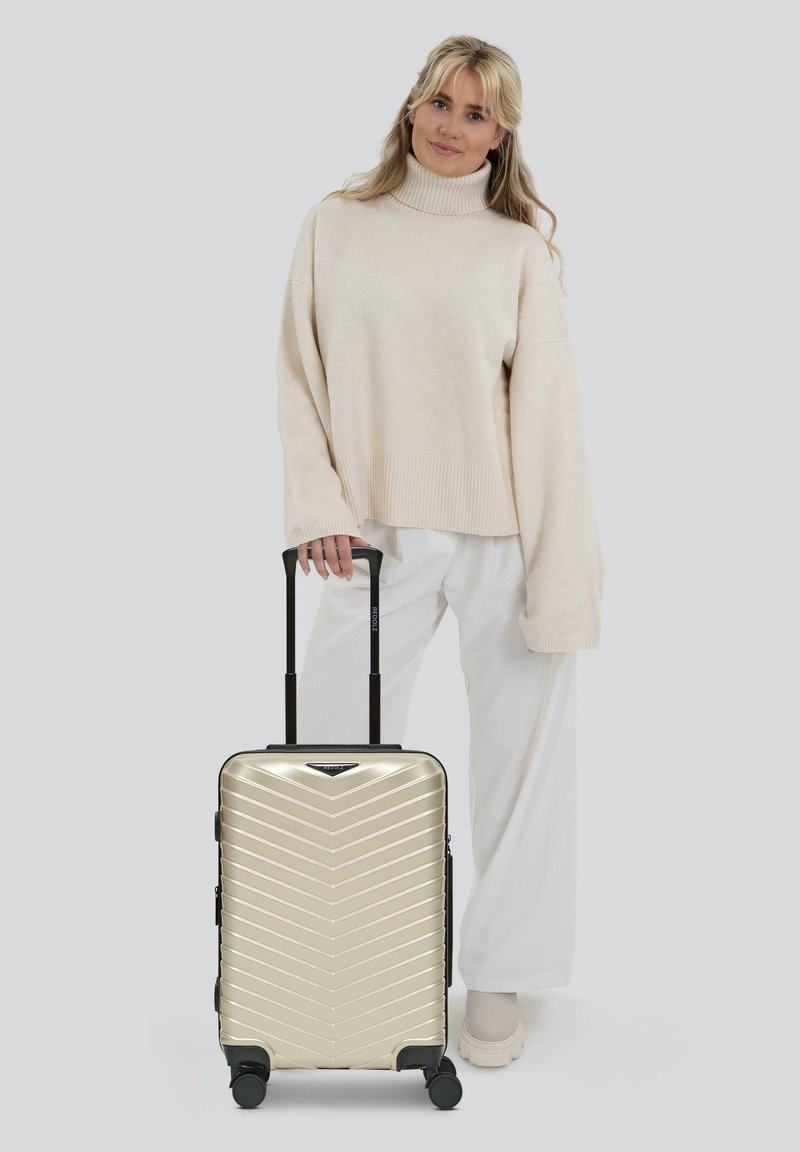 Goldfarbener Hartschalenkoffer mit Chevron-Muster, schwarzen Akzenten, ausziehbarem Griff und 4 Rädern, präsentiert neben einer Person in einem cremfarbenen Pullover und weißen Hosen.