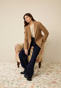 Bruine wollen blazer met bloemrijke borduursels, crèmekleurige gebreide top en donkerblauwe flare jeans. Zwarte schoenen maken de outfit compleet. Achtergrond met bloemenpatroon.