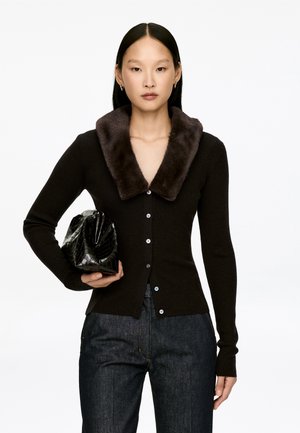 Dunkelbraune gerippte Strickjacke mit einem Kunstfellkragen, die vorne mit Knöpfen versehen ist. Das Model hält eine schwarze, strukturierte Handtasche.