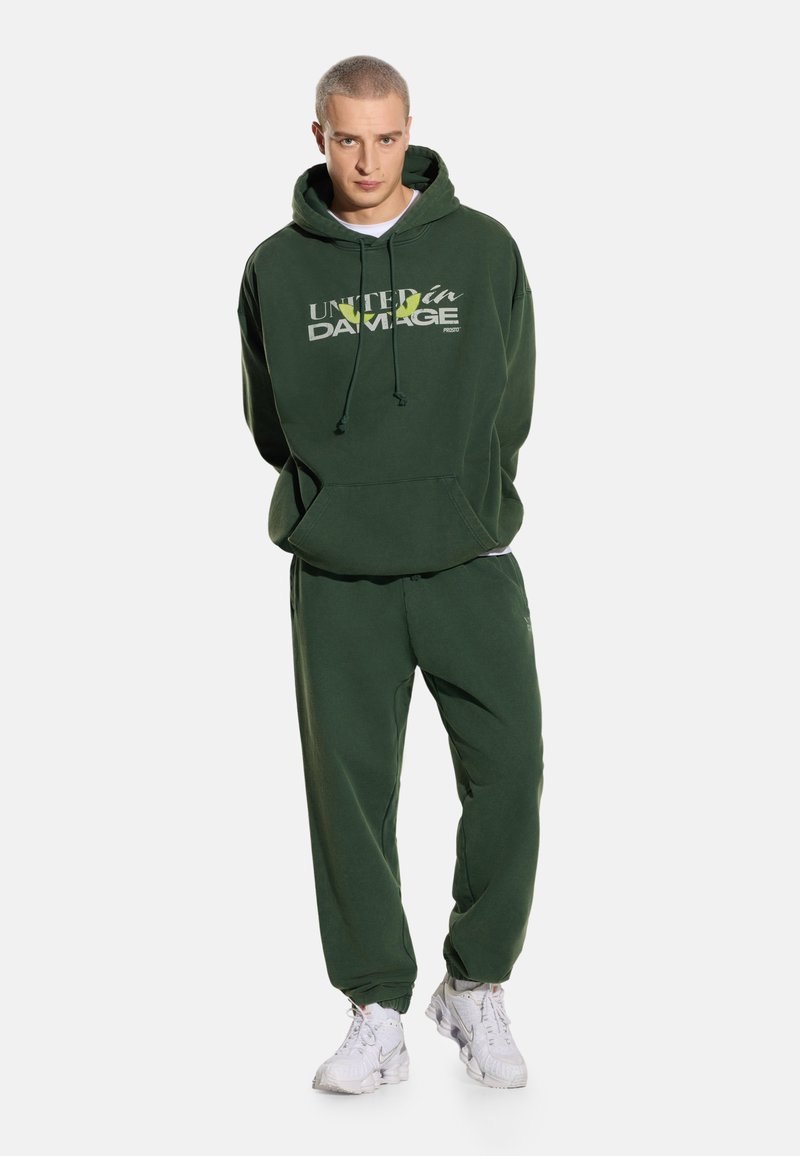 Mężczyzna w ciemnozielonej bluzie z kapturem z napisem „United in Damage” i dopasowanych spodniach dresowych, stojący z rękami za plecami na białym tle.