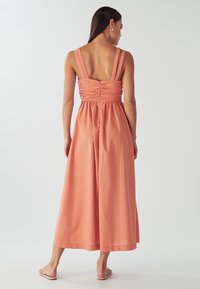 Virsikuvärvi maksikleit paksude rihmadega, seljal kogutud kangaga ja lehviva A-lõikega seelikuga. Sile tekstuur ja vabaaja disain.