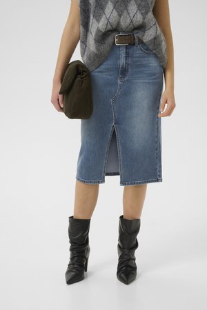 Woman wearing a blue denim skirt with front slit, black heeled boots, gray sleeveless sweater, and holding a dark green clutch bag.