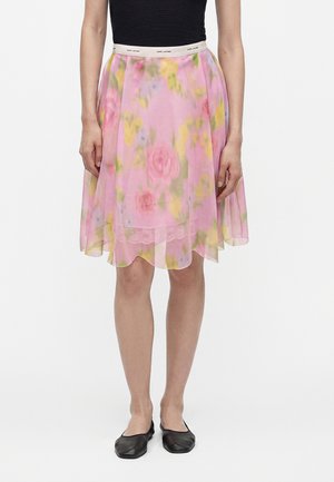 Personne portant une jupe rose à fleurs, longueur aux genoux, avec une ceinture blanche, un haut noir sans manches et des ballerines noires, debout bien droite.