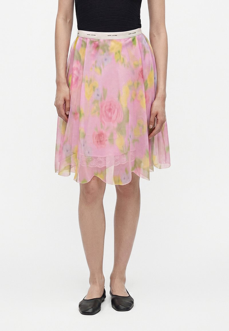 Personne portant une jupe rose à fleurs, longueur aux genoux, avec une ceinture blanche, un haut noir sans manches et des ballerines noires, debout bien droite.