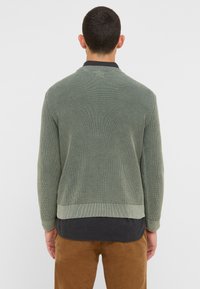 Pull en maille côtelée verte avec un col rond, surface texturée et taille ajustée, superposé sur une chemise sombre et un pantalon beige.