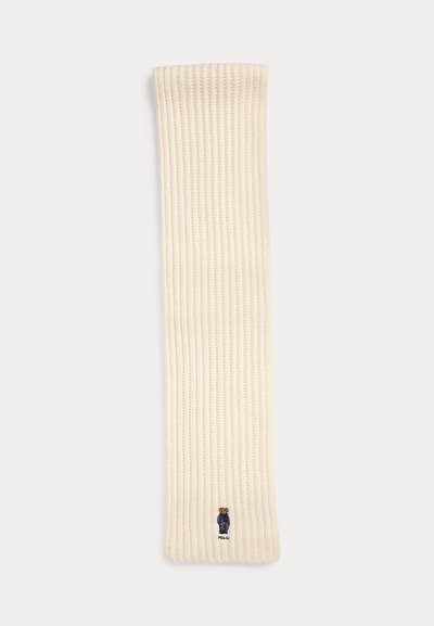 Kremna rebrasta pletena ruta z izvezenim medvedjim logotipom na dnu. Mehka tekstura s pravokotno obliko, približno 150 cm dolžine.