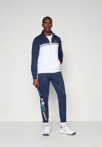 Marineblå og hvit treningsdress laget av lett stoff. Jakken har høy krage og glidelås, buksene har elastisk midje. Tennisracket holdt i én hånd.