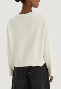 Pull en maille couleur crème avec un ourlet et des poignets côtelés, présentant une coupe décontractée. Associé à un jean denim noir avec des poches arrière visibles.