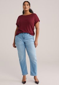 Bordeauxfarbenes Kurzarmoberteil mit strukturiertem Zickzackmuster, kombiniert mit hellblauen Jeans mit geradem Bein und dunklen, spitzen Absätzen.