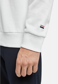 Main d'homme reposant sur le côté portant un sweat-shirt blanc avec un logo rayé rouge, blanc et bleu sur le revers de la manche.