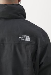 Giacca nera con colletto alto, realizzata in un tessuto liscio. Presenta un logo bianco ricamato sulla schiena, posizionato sulla spalla.