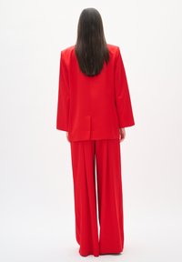 Traje rojo a medida con un blazer largo y pantalones de pierna ancha. Tejido suave y texturizado; ligero brillo; líneas limpias y diseño minimalista.