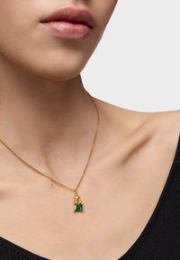 Zbliżenie na szyję osoby noszącej złoty naszyjnik z małym zielonym prostokątnym kamieniem wisiorkiem, ubraną w czarną bluzkę.