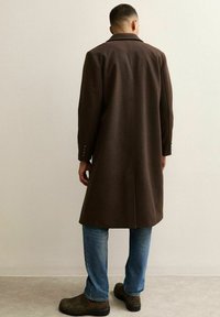 Abrigo de lana marrón con un corte entallado, abertura trasera sencilla y puños abotonados, combinado con jeans de un azul claro y calzado oscuro.