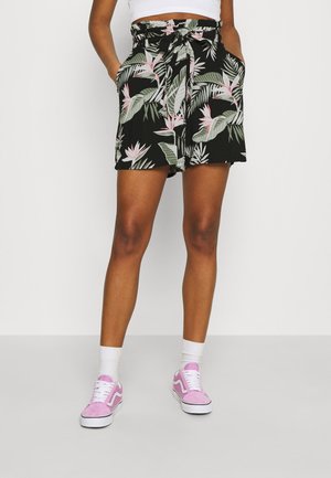 Kvinde iført sorte paperbag taljeshorts med grønne og pink tropiske bladmønstre, hvide sokker og pink sneakers, står med hænderne i lommerne.