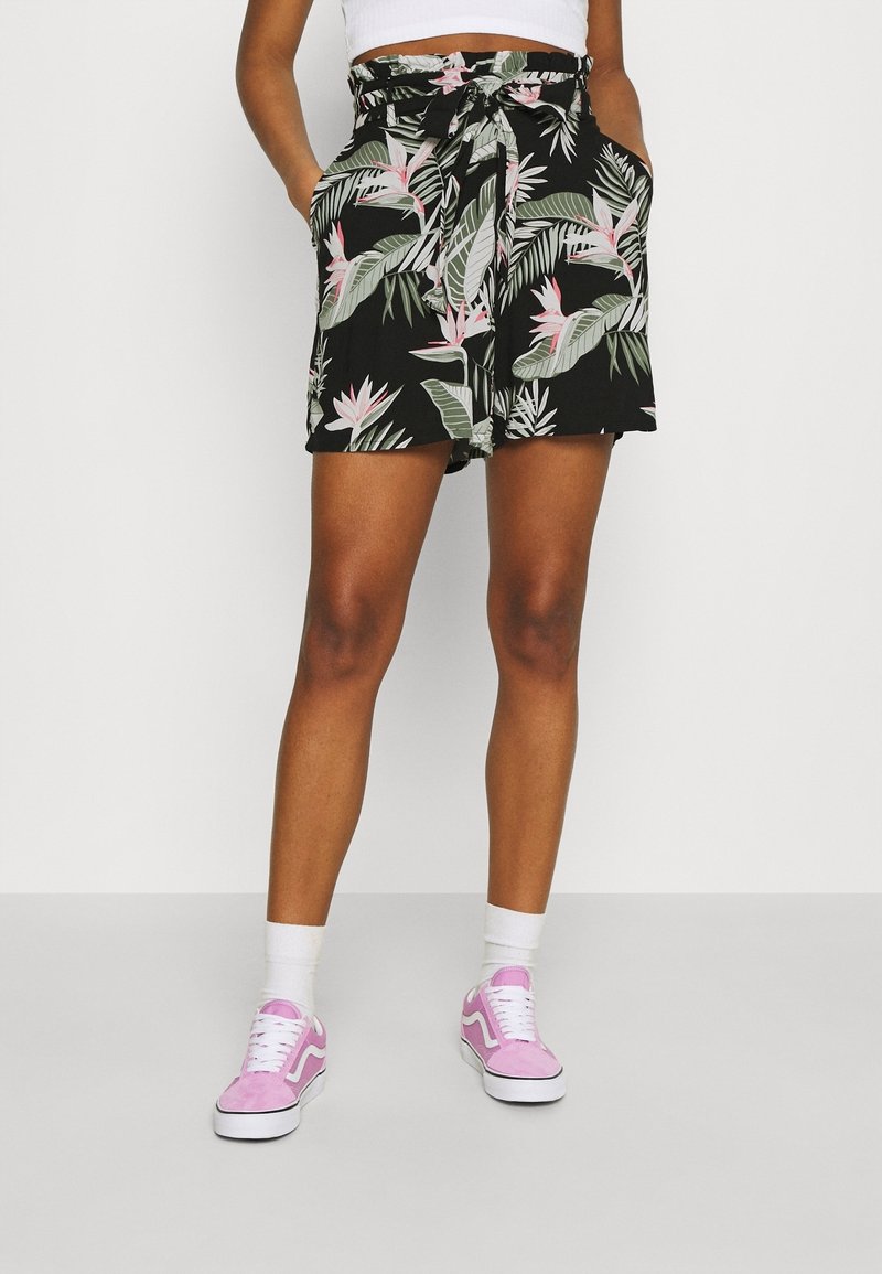 Femme portant un short taille paperbag noir avec un motif de feuilles tropicales vertes et roses, des chaussettes blanches et des baskets roses, debout les mains dans les poches.
