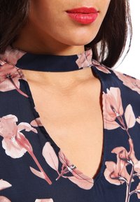 Blusa con stampa floreale caratterizzata da un profondo scollo a V e un colletto aderente in stile choker. Realizzata in tessuto leggero di colore blu navy con motivi floreali rosa.