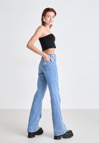 Jeune femme debout, portant un crop top noir sans bretelles, un jean taille haute bleu clair à jambes larges et des bottes noires à semelle épaisse.
