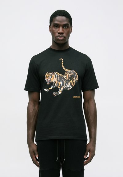 Homme portant un t-shirt noir avec un graphisme de tigre orange et blanc et le petit texte "roberto cavalli" sur le devant, se tenant contre un fond uni.
