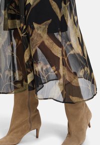 Braune Wildleder-Stiefel mit Absatz unter einem transparenten schwarzen Rock mit einem großen goldenen und braunen Blattmuster.
