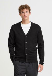 Svart cardigan laget av mykt materiale, med V-hals, knappelukking og ribbede ermer. Båret over en hvit skjorte, med et enkelt design.
