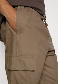 Cargo kalhoty v tlumené hnědé látce. Mají elastický pas, boční kapsy a klopovou kapsu, což zajišťuje hladkou texturu.