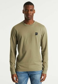 Camicia a maniche lunghe verde oliva realizzata in morbido tessuto, con una toppa nera con logo sul petto, scollatura rotonda e polsini aderenti.