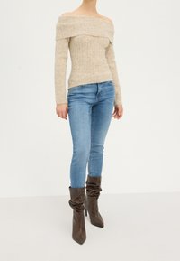 Beige off-shoulder gebreide trui met ribbelstructuur, gecombineerd met lichtblauwe slim-fit jeans en bruine slouchy haklaarzen.