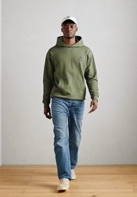 Olivovo zelená mikina s kapucňou a klokaním vreckom, v kombinácii so svetlo modrými džínsami a svetlými teniskami. Biela čiapka s detailom loga.