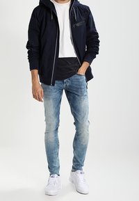 Navy hooded jacket with zipper pocket, white t-shirt underneath, light blue distressed skinny jeans, and white sneakers.