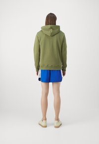 Olivově zelená mikina s kapucí, texturovaná tkanina, spárovaná s modrými šortkami. Jedinec má na sobě světlé boty a stojí proti bílému pozadí.