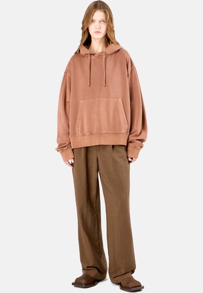Ontspannen hoodie in stofroze met een kangoeroezak, een capuchon met trekkoorden en een dropped shoulder, gecombineerd met losse bruine broek en bruine schoenen.