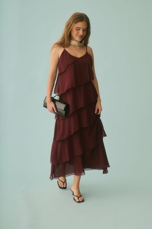 Nő réteges bordó maxiruhában és fekete szandálban, fekete táskát viselve, egyszínű világoskék háttér előtt sétál.