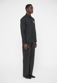 Camisa negra de manga larga con diseño abotonado y logo circular dorado en el pecho izquierdo. Combinada con pantalones anchos gris oscuro.