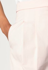 Calças de algodão cor-de-rosa claro com pregas, uma textura suave e um corte ajustado. Apresenta bolsos laterais e detalhes na cintura.