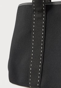 Μαύρη τσάντα tote από υφασμάτινη ύλη, με φαρδιές μαύρες ραμμένες λαβές και λευκή ραφή κατά μήκος των ακμών, σε απλό φόντο.