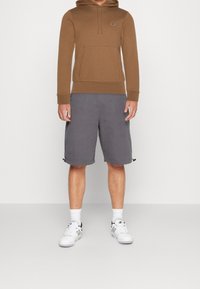 Brown hoodie with a front pocket, paired with gray shorts. White sneakers with black accents and visible socks. Simple and casual design.