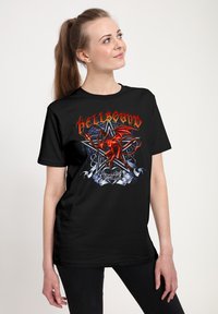 Žena s dlouhým culíkem, oblečená v černém tričku s červeným ďáblem, pentagramem, trny a textem "Hellbound" napsaným ohnivým písmem.