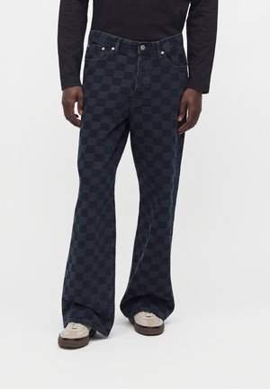 Homme portant un pantalon large à carreaux noir et gris foncé, une chemise noire à manches longues et des baskets beiges sur fond blanc uni.