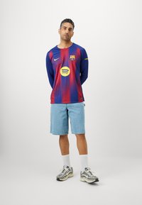 Dugout FC Barcelone dugoročni rukav majica s gradijentom plave i crvene boje, s žutim Spotify logotipom i grbom kluba, nosi se uz traper šorc i tenisice.