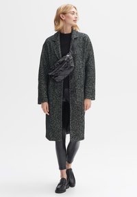 Long, dark gray coat with a textured, herringbone pattern. Paired with black leggings and chunky loafers. Features a shiny black crossbody bag.