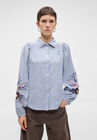 Ριγέ μπλε και λευκό πουκάμισο με κουμπιά και φουσκω τά μανίκια; διαθέτει λουλούδια κεντημένα στους καρπούς σε αποχρώσεις ροζ, μωβ και καφέ.