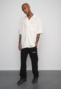Camisa corta de manga corta color crema claro con un bolsillo, combinada con pantalones de chándal negros que tienen texto blanco. Lleva puesto unos zapatos deportivos negros.