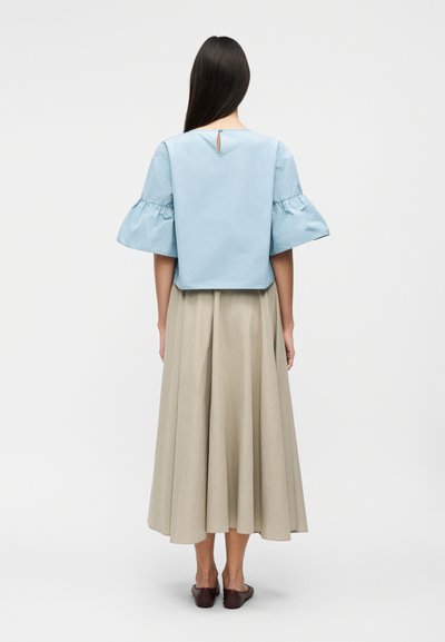 Femme aux longs cheveux foncés portant un chemisier bleu clair à manches courtes avec des volants aux manches et une jupe midi beige, debout de dos.