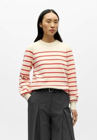 Maglione crema con strisce orizzontali rosse, scollo rotondo e maniche a sbuffo, abbinato a pantaloni grigi plissettati a vita alta e cintura nera.