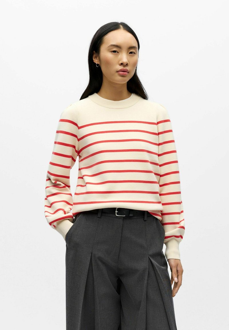 Maglione crema con strisce orizzontali rosse, scollo rotondo e maniche a sbuffo, abbinato a pantaloni grigi plissettati a vita alta e cintura nera.