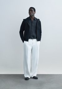 Ein Mann steht mit den Händen in den Taschen, trägt einen schwarzen Kapuzenpullover mit Reißverschluss, ein schwarzes Hemd, weiße, weit geschnittene Hose und schwarze Schuhe vor einem schlichten Hintergrund.