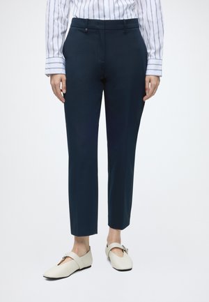 Donna che indossa pantaloni blu navy su misura, una camicia bianca e blu a righe e scarpe basse panna con fibbie.