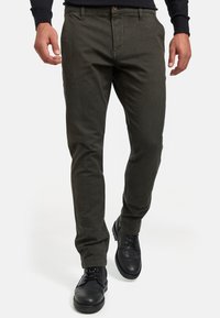 Pantalones ajustados de color verde oscuro hechos de tejido texturizado, con cremallera en la parte delantera, bolsillos laterales y zapatos de vestir de cuero negro.
